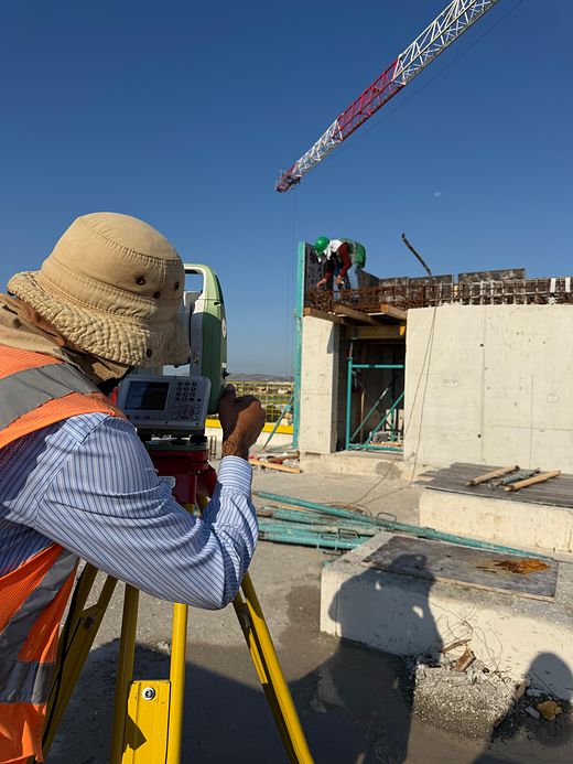 An ORIGIN surveyor in a high-visibility vest sighting through a Leica total station on a construction site in Malta, with a tower crane and reinforced-concrete formwork behind him.
