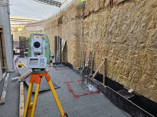 Total station on a tripod beside an excavated rock face, with red setting-out marks on the slab and reinforcement starter bars.
