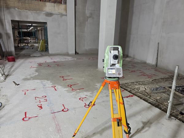 A Leica total station set up on a tripod on a concrete slab, with red setting-out marks and a dimension note written on the floor.