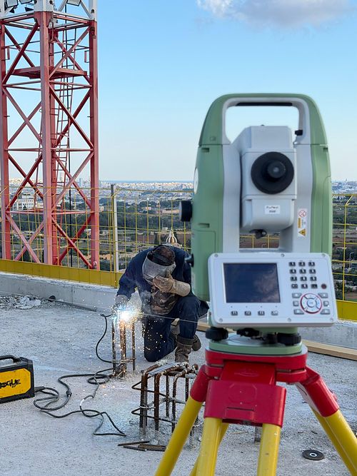 A Leica total station on a tripod on a rooftop slab, with a worker welding reinforcement starter bars behind it and the Maltese skyline in the distance.