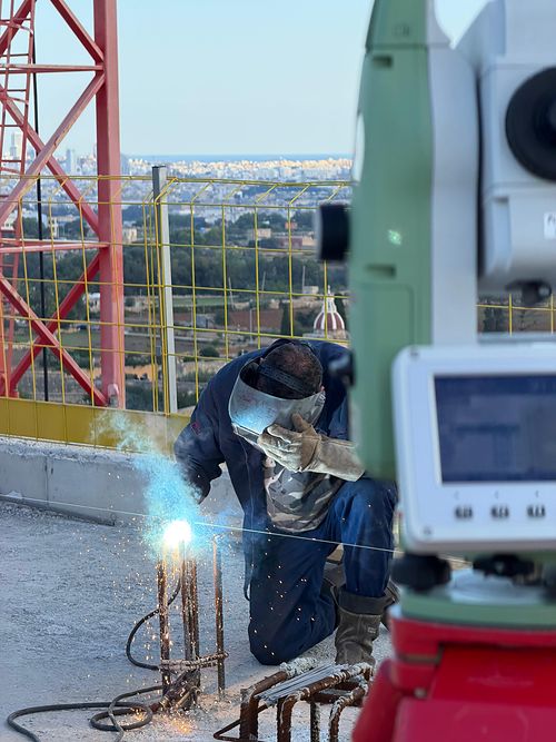 A worker in a welding mask joining reinforcement starter bars beside a total station, with a tower crane mast and the Maltese skyline behind.