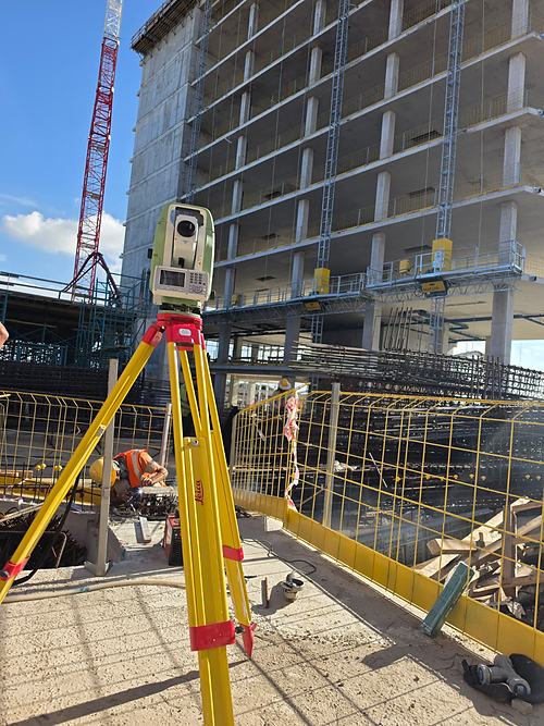 A Leica total station on a yellow tripod on a concrete slab, with reinforcement cages, a tower crane and a multi-storey concrete frame under construction behind it.